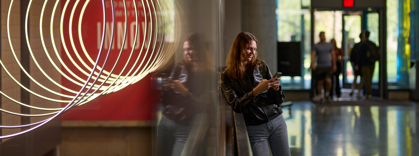 Female Iowa State student looking at cell phone and leaning against wall. Doorway with students walking in the background and red wall art with ring lights.